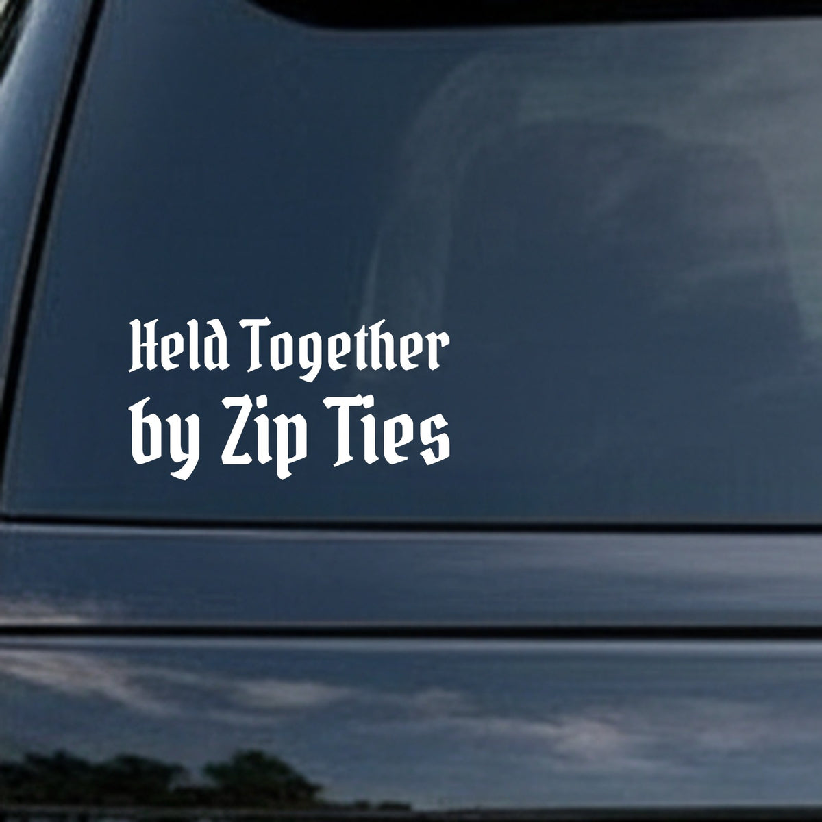 White gothic Old English style lettering stating Held Together by Zip Ties on a black background for a car decal