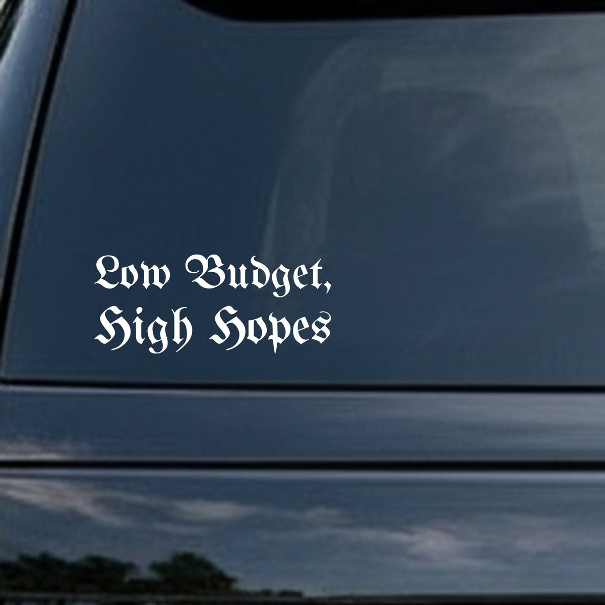 White vinyl decal reading Low Budget, High Hopes in Gothic Old English style font on black background