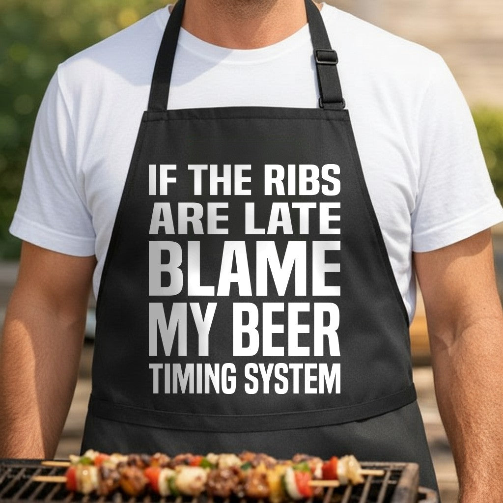 Black apron with large white bold text saying If The Ribs Are Late Blame My Beer Timing System in a clean typography style.