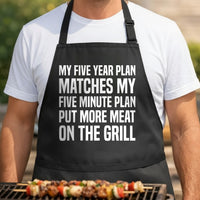 Black apron with bold white text saying My Five Year Plan Matches My Five Minute Plan Put More Meat On The Grill in a block font.