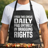 Black apron with large white block text reading I feed this circus daily paid entirely in bragging rights in bold typography