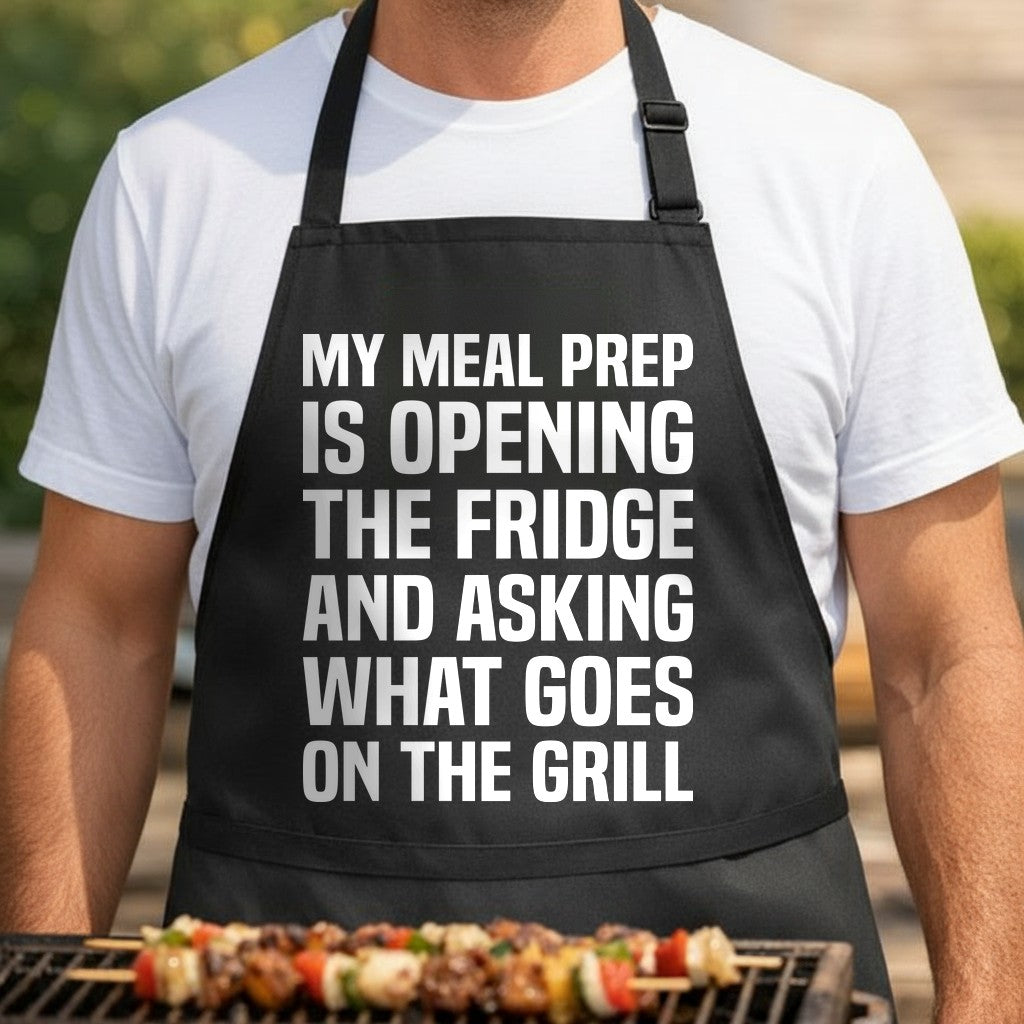 Black apron with large white text reading My meal prep is opening the fridge and asking what goes on the grill in bold uppercase letters