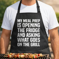 Black apron with large white text reading My meal prep is opening the fridge and asking what goes on the grill in bold uppercase letters