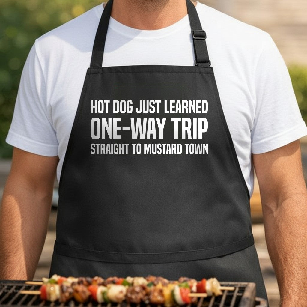 Black apron with large white text reading hot dog just learned one-way trip straight to mustard town in bold font