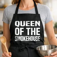 Black BBQ apron with large white text reading Queen of the Smokehouse in bold, uppercase letters.