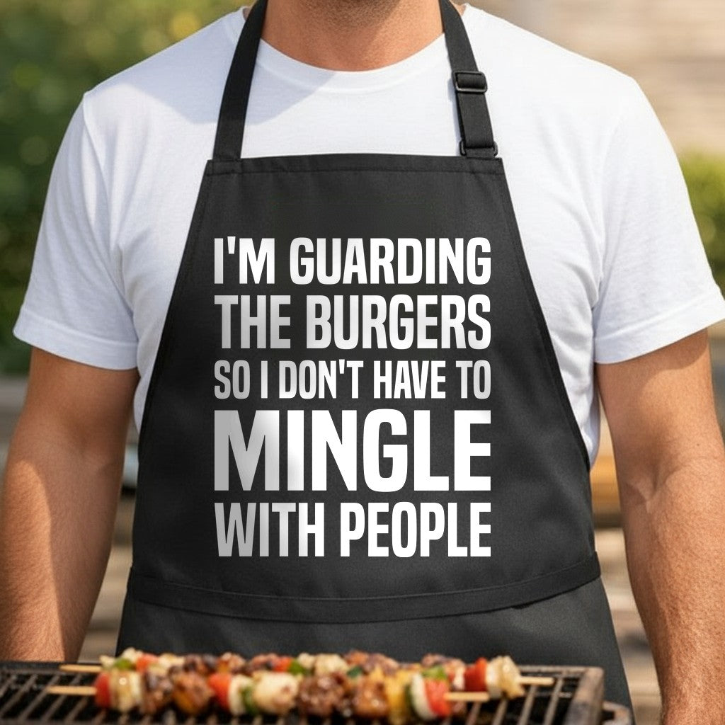 Black apron with large white bold text reading I’m guarding the burgers so I don’t have to mingle in a simple block font
