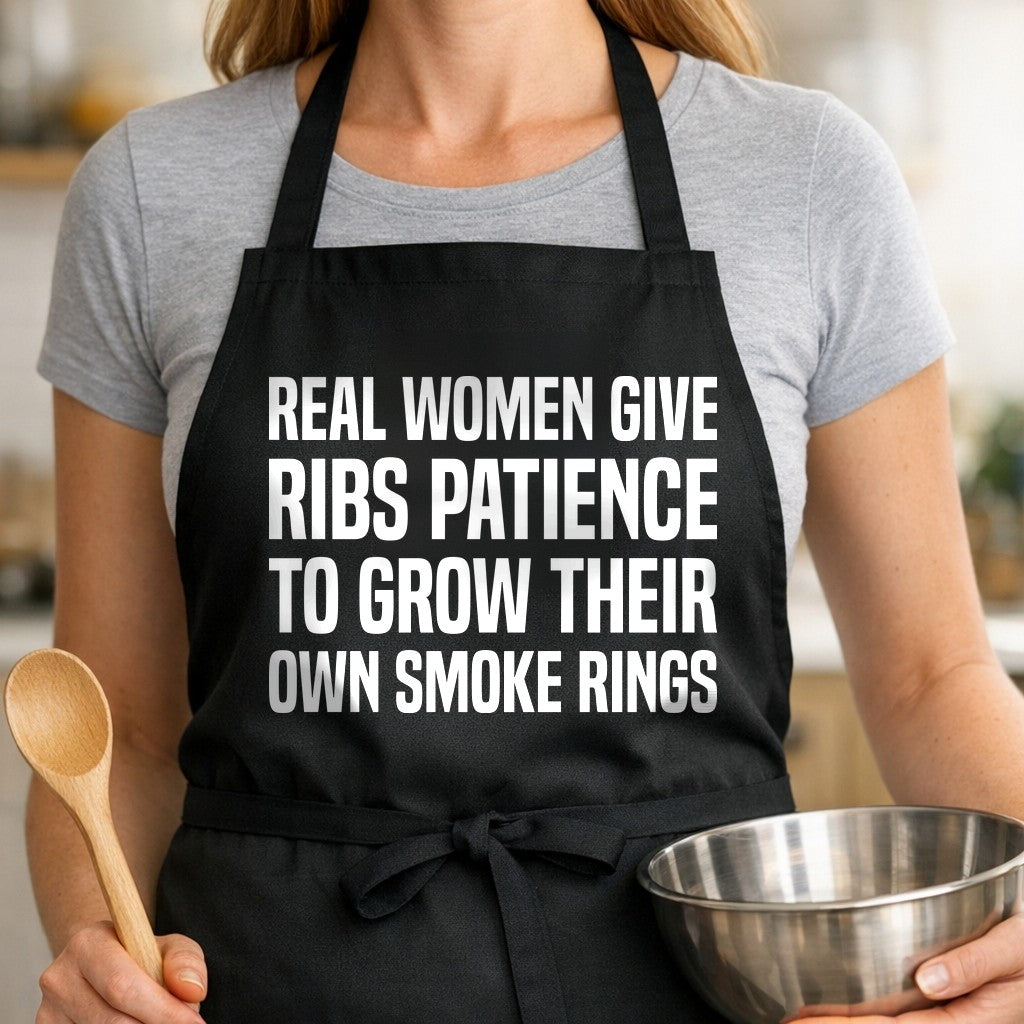 Black apron with large white text reading Real Women Give Ribs Patience To Grow Their Own Smoke Rings in bold block letters