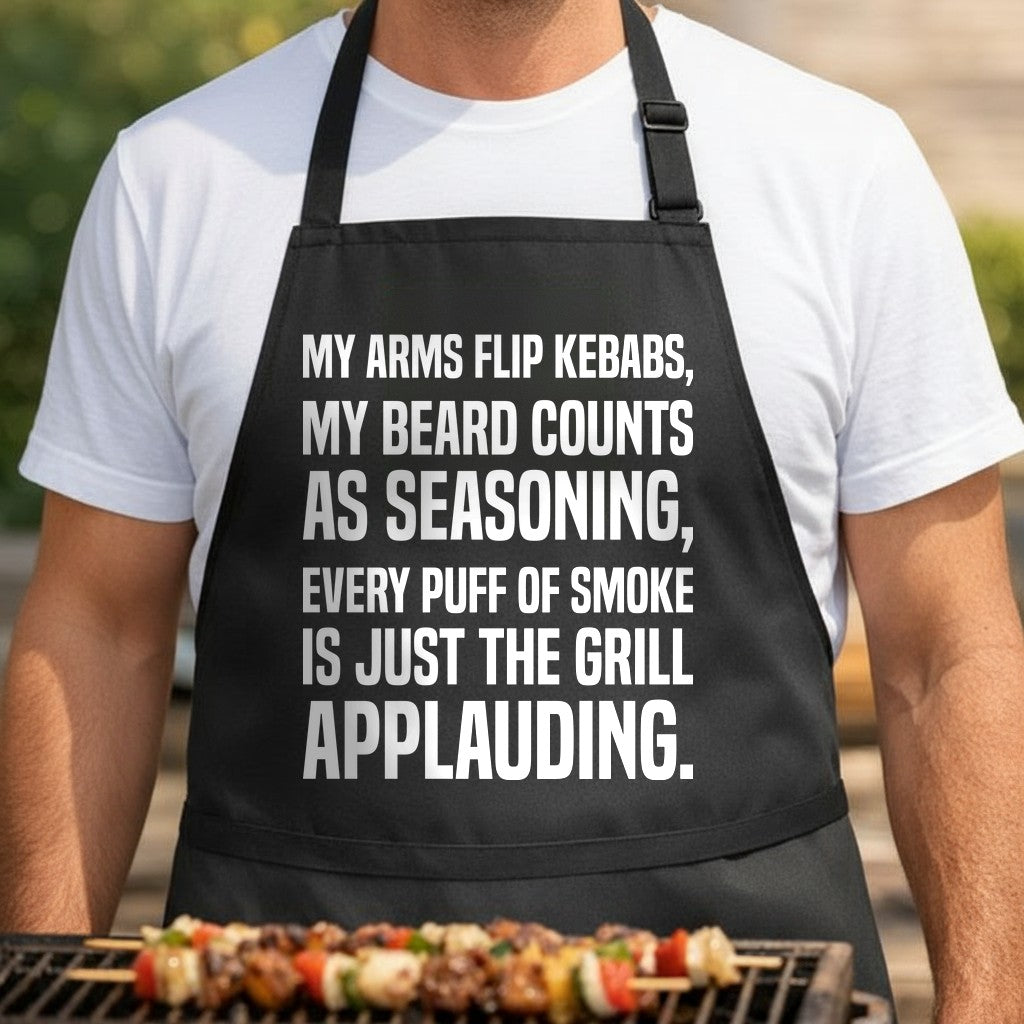 Black BBQ apron with bold white text humorously praising grilling skills, referencing arms, beard, and smoke applause.