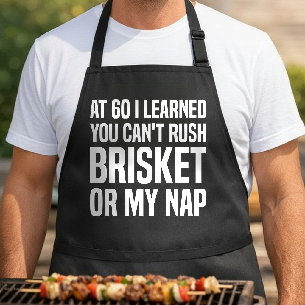 Black apron with large white block text saying At 60 I Learned You Can't Rush Brisket Or My Nap in bold uppercase letters