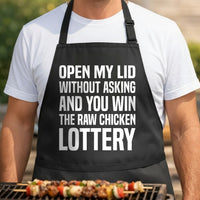 Black apron with large white text reading open my lid without asking and you win the raw chicken lottery in bold font