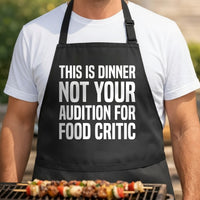 Black apron with large white bold text stating This Is Dinner Not Your Audition For Food Critic in a simple block font