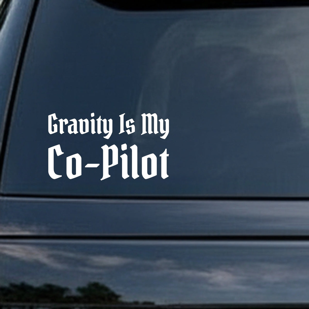 White vinyl decal displaying the phrase Gravity Is My Co-Pilot in gothic blackletter font on a black background.