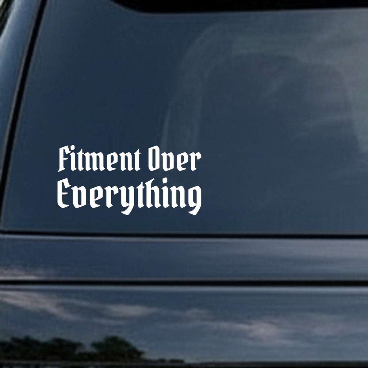 White vinyl decal with the phrase Fitment Over Everything in gothic blackletter Old English font on a black background.