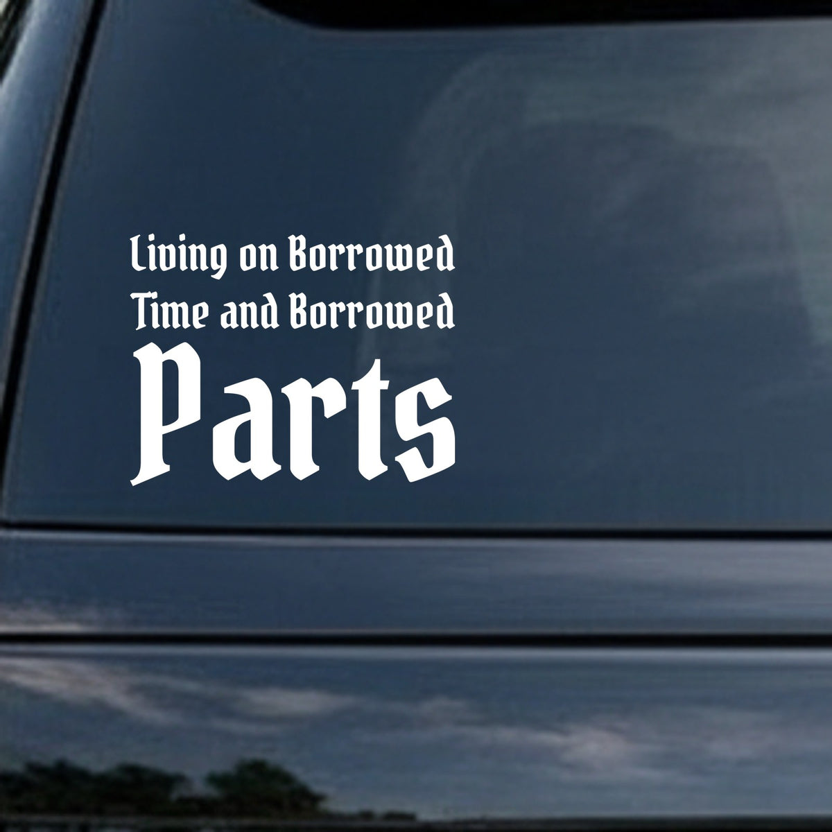 White vinyl decal with the phrase Living On Borrowed Time And Borrowed Parts in gothic blackletter Old English font on a black background.