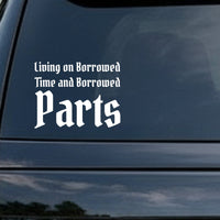 White vinyl decal with the phrase Living On Borrowed Time And Borrowed Parts in gothic blackletter Old English font on a black background.