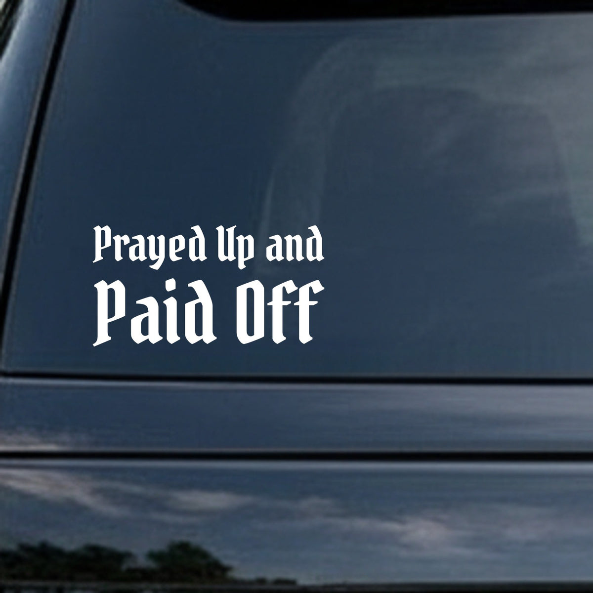 White vinyl car decal with the phrase 'Prayed Up and Paid Off' in gothic blackletter style on a black background