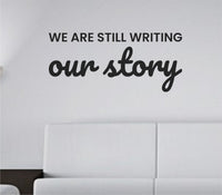 Black vinyl wall decal displaying the phrase 'We are still writing our story' in a simple modern font on a white background.