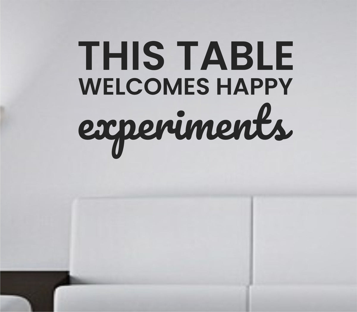 Black vinyl wall decal with the phrase 'This table welcomes happy experiments' in a simple, friendly font on a white background.