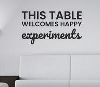 Black vinyl wall decal with the phrase 'This table welcomes happy experiments' in a simple, friendly font on a white background.