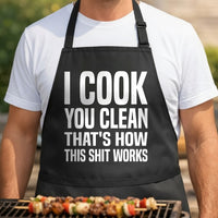 Black apron with large white bold text reading I Cook You Clean That's How This Shit Works in a block style font.
