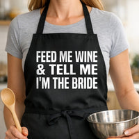 Black apron with large white text reading Feed Me Wine And Tell Me I'm The Bride in bold block letters, simple and bold style.