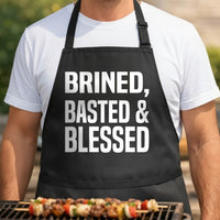 Black apron with large white bold text reading 'Brined, Basted & Blessed' in a simple block style.