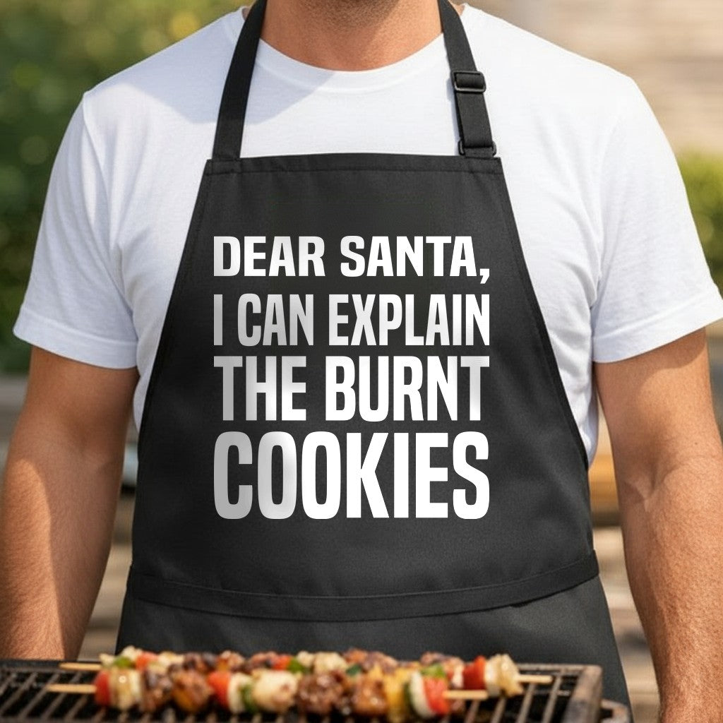 Black apron with large white text saying 'Dear Santa I Can Explain The Burnt Cookies' in a simple bold font.