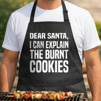 Black apron with large white text saying 'Dear Santa I Can Explain The Burnt Cookies' in a simple bold font.