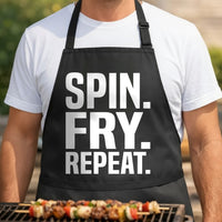 Black apron with large white text reading Spin Fry Repeat in bold letters, simple graphic design style