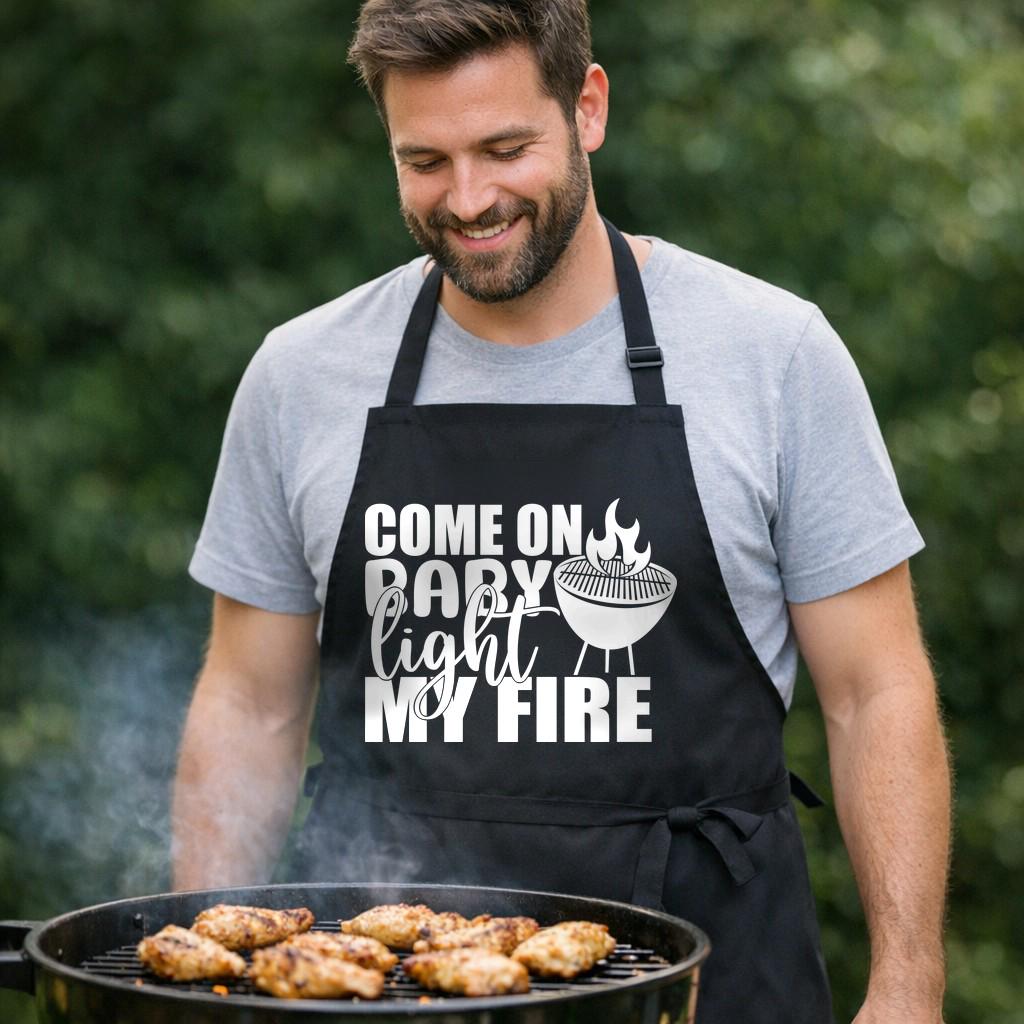 Come On Baby Light My Fire black BBQ apron, at the grill: Black apron with white text saying Come On Baby Light My Fire and a graphic of a round grill with flames on top