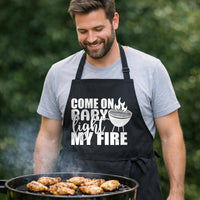 Come On Baby Light My Fire black BBQ apron, at the grill: Black apron with white text saying Come On Baby Light My Fire and a graphic of a round grill with flames on top