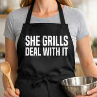 She Grills, Deal With It black BBQ apron, woman torso close-up: Black apron with bold white uppercase text reading 'SHE GRILLS DEAL WITH IT' in a simple typographic design