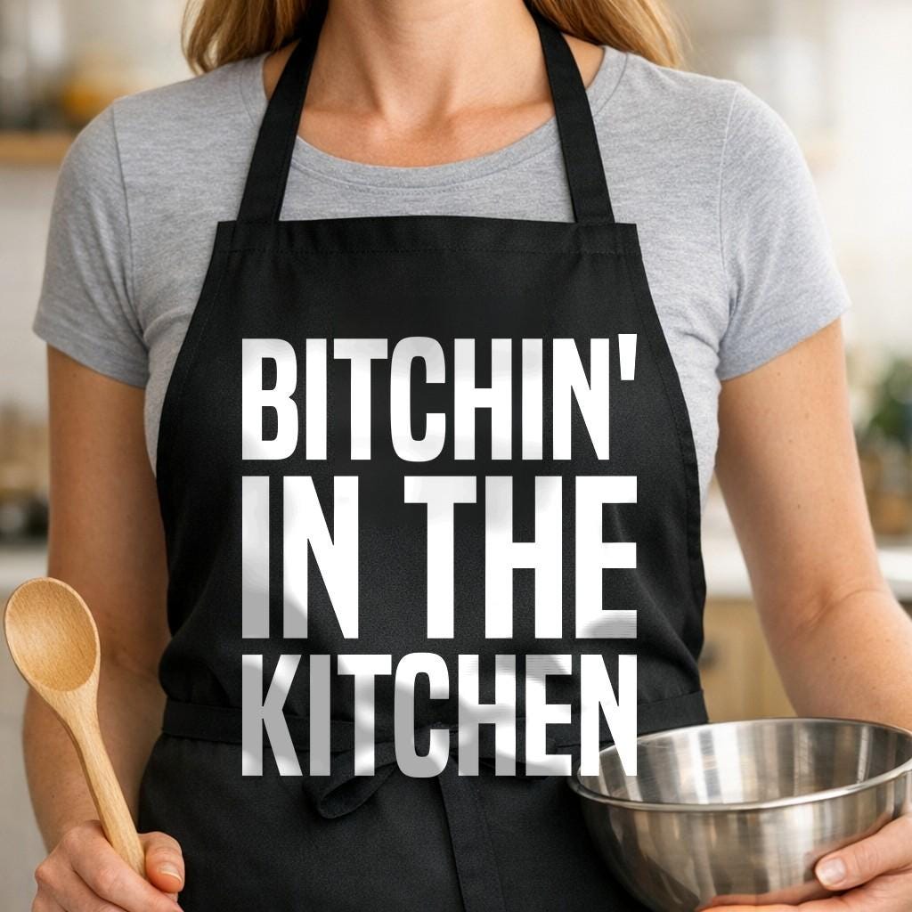 Bitchin' in the Kitchen black BBQ apron, woman torso close-up: Black apron with large white bold text reading Bitchin' in the Kitchen in a blocky uppercase font