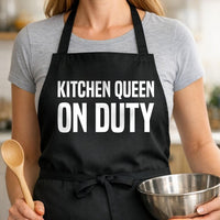 Kitchen Queen On Duty black BBQ apron, woman torso close-up: Black apron with large white bold text reading Kitchen Queen On Duty in simple typography on solid black background