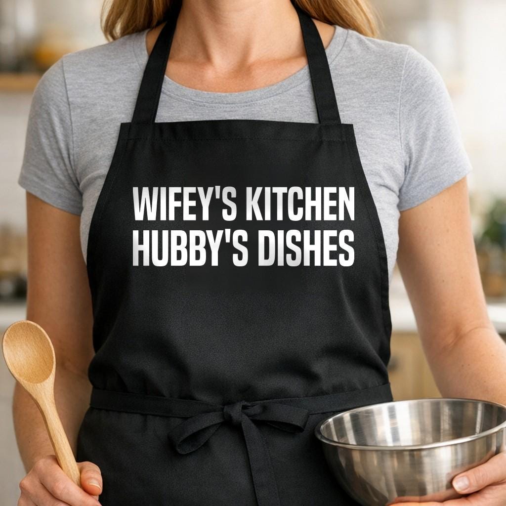 Wifey's Kitchen, Hubby's Dishes black BBQ apron, woman torso close-up: Black apron with bold white uppercase text reading 'Wifey's Kitchen Hubby's Dishes' in a clean, modern font