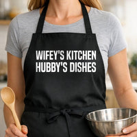 Wifey's Kitchen, Hubby's Dishes black BBQ apron, woman torso close-up: Black apron with bold white uppercase text reading 'Wifey's Kitchen Hubby's Dishes' in a clean, modern font