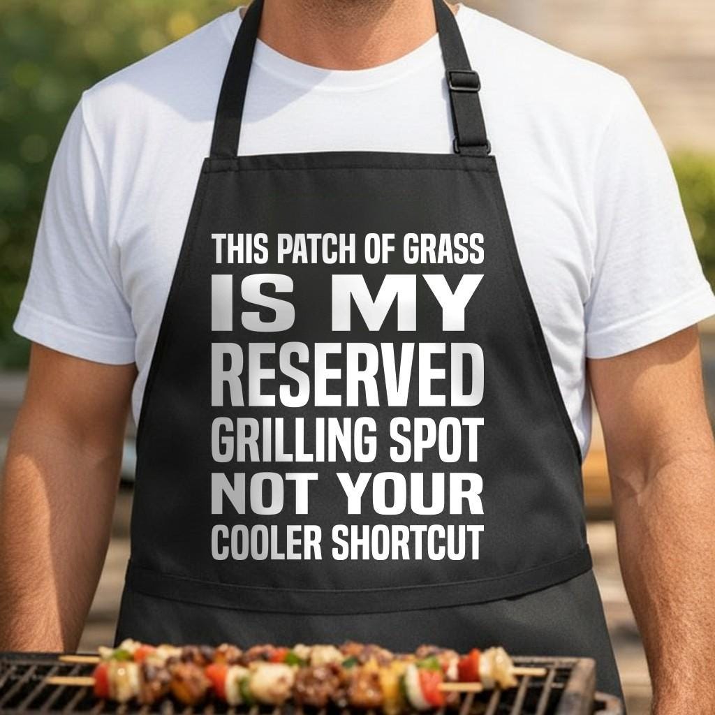 Reserved Grill Spot black BBQ apron, torso close-up: Black apron with large white bold text stating a reserved grilling spot is marked by a worn patch of grass, humorous barbecue message.