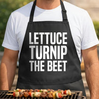 Lettuce Turnip the Beet black BBQ apron, torso close-up: Black apron with large white text saying Lettuce Turnip The Beet in bold block letters, simple graphic design.
