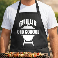 Grillin' Old School black BBQ apron, torso close-up: Black apron with white graphic showing a kettle grill silhouette and the words 'Grillin' Old School' in bold letters.