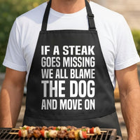 Blame The Dog black BBQ apron, torso close-up: Black apron with large bold white text stating If a steak goes missing we all blame the dog and move on in a simple font