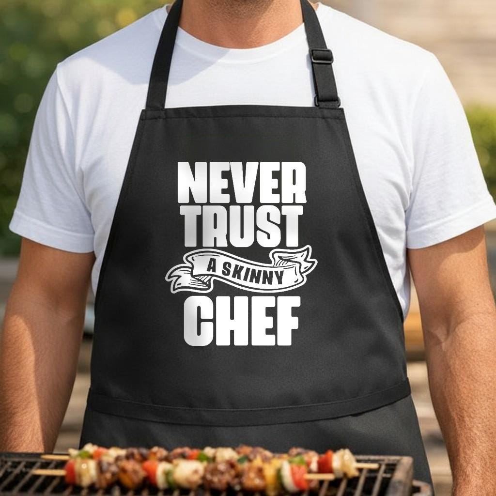 Never Trust a Skinny Chef black BBQ apron, torso close-up: Black apron with large white bold text reading Never Trust a Skinny Chef with a banner design around the word skinny