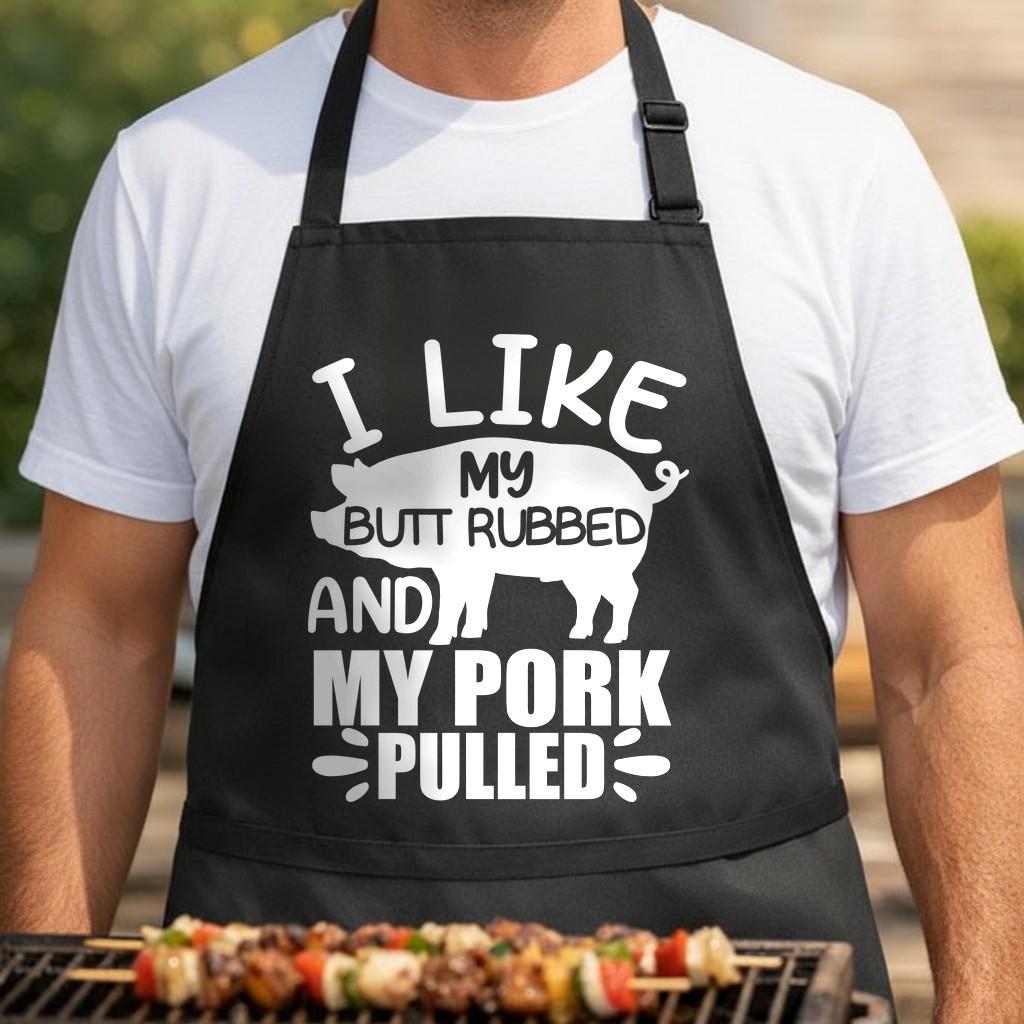 I Like My Butt Rubbed and My Pork Pulled black BBQ apron, torso close-up: Black apron with white artwork showing a pig silhouette and text reading I Like My Butt Rubbed And My Pork Pulled in bold letters