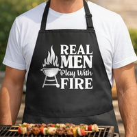 Real Men Play with Fire black BBQ apron, torso close-up: Black apron with white text reading Real Men Play With Fire and an illustration of a flaming barbecue grill on the left