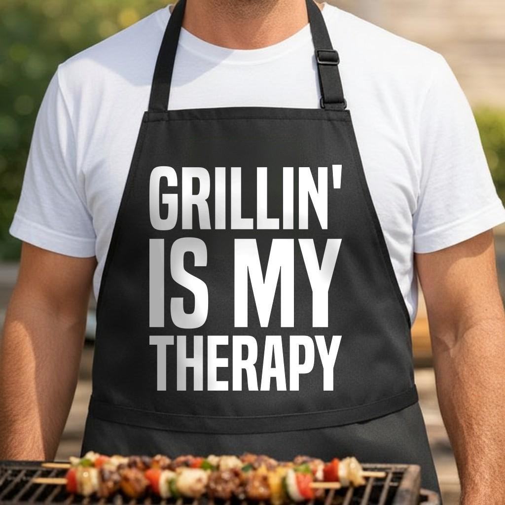 Grillin' Is My Therapy black BBQ apron, torso close-up: Black apron with large white bold text saying Grillin' Is My Therapy in a stacked layout