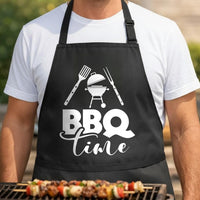 BBQ Time black BBQ apron, torso close-up: Black apron with white design showing a kettle grill with lid open, spatula and fork crossed above, and the words BBQ Time below in bold and script fonts.