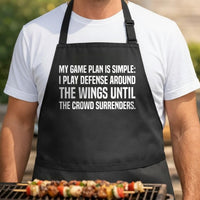 Snack Defense Captain black BBQ apron, torso close-up: Black apron with large white text reading a humorous grilling message about playing defense around chicken wings at a barbecue.