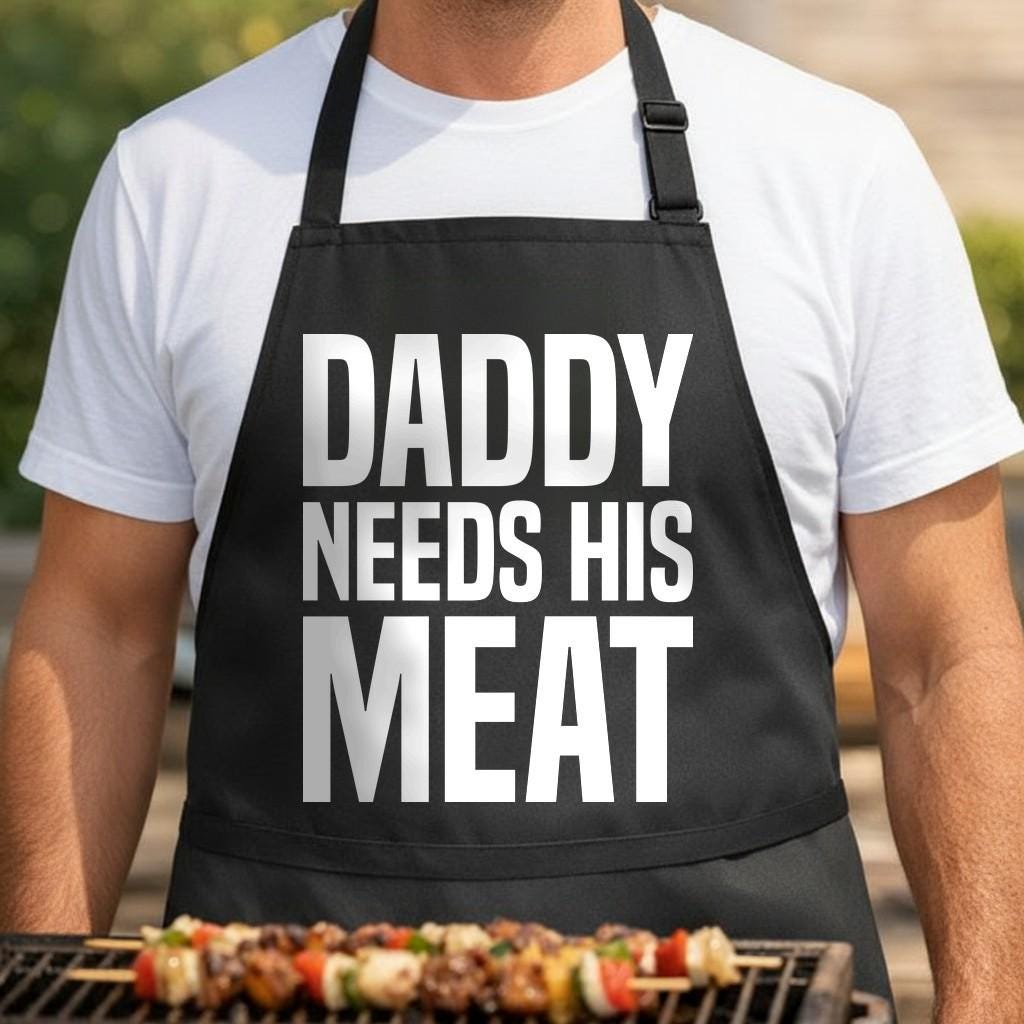Daddy Needs His Meat black BBQ apron, torso close-up: Black apron with large white block letters reading Daddy Needs His Meat in bold typography style
