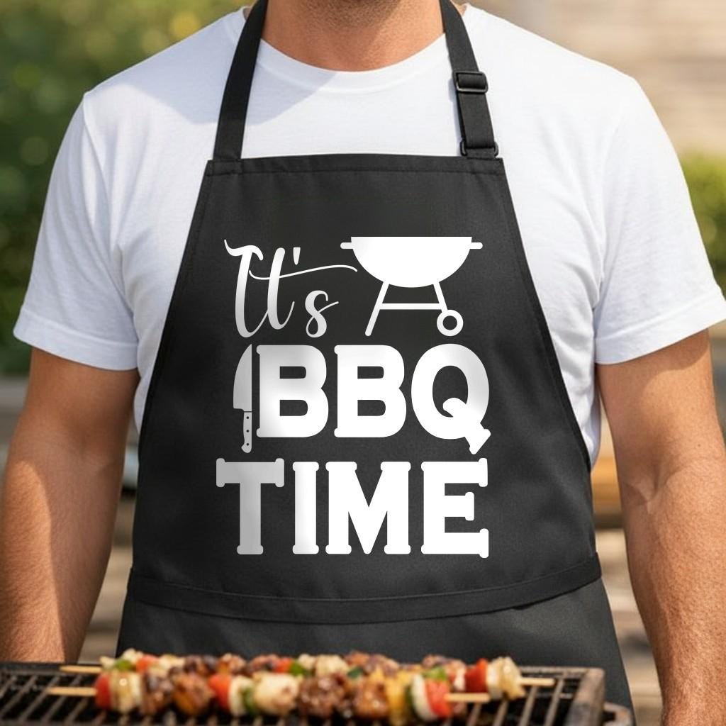 It's BBQ Time black BBQ apron, torso close-up: Black apron with white graphic text 'It's BBQ Time' including a kettle grill, knife, and spatula icons in bold, simple design.