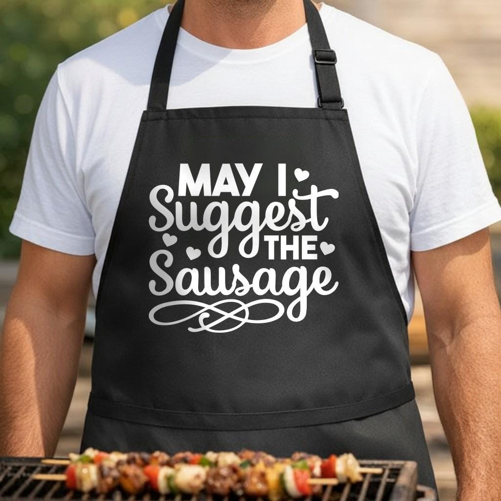 May I Suggest the Sausage black BBQ apron, torso close-up: Black BBQ apron with white text reading 'May I Suggest The Sausage' in mixed fonts with small heart decorations and a decorative swirl.