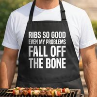 Bone-Deep Therapy black BBQ apron, torso close-up: Black apron with large white block text reading ribs so good even my problems fall off the bone in a bold uppercase font